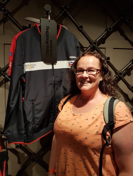 Blind veteran Clare smiling, stood next to her Invictus Games jacket which is hung up behind her