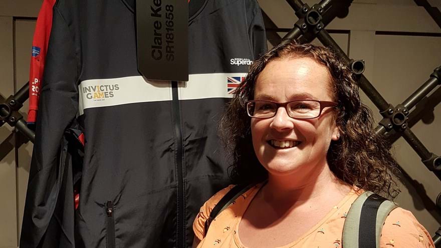 Blind veteran Clare smiling, stood next to her Invictus Games jacket which is hung up behind her
