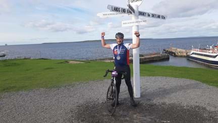 Jon stood under a signpost for John O'Groats with his arms in the air and medal around his neck
