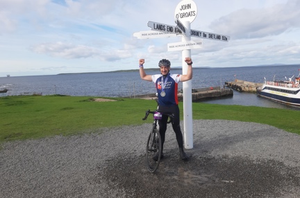 Jon stood under a signpost for John O'Groats with his arms in the air and medal around his neck