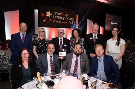 A group of Blind Veterans UK staff, sitting and standing at a round table at an awards ceremony, with a banner behind them that reads: Smee & Ford Legacy Giving Awards 2023