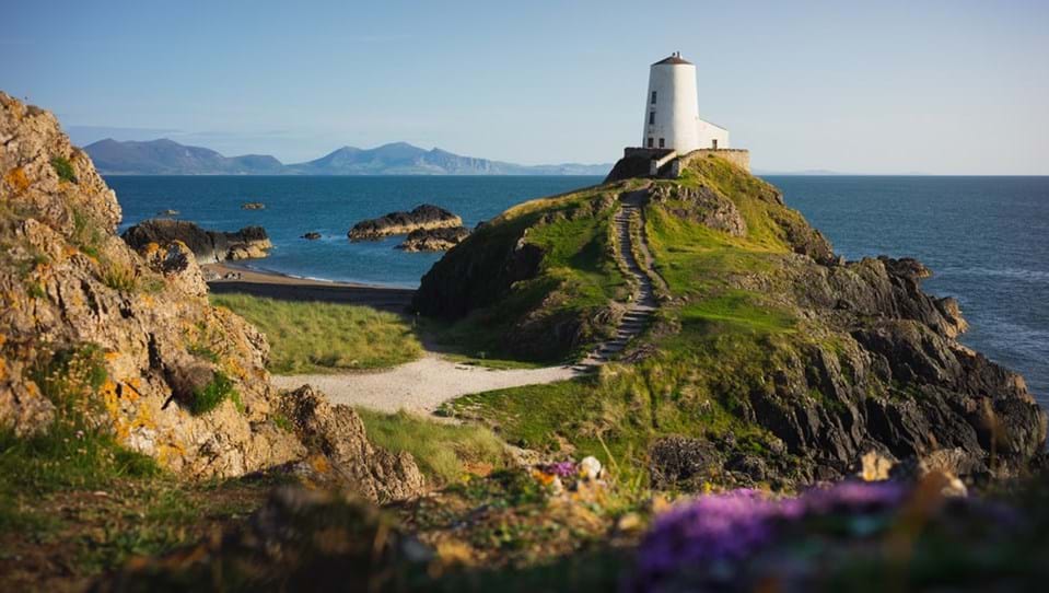 The Twr Mawr Lighthouse placed upon a green hill on a sandy beach, with mountains in the background