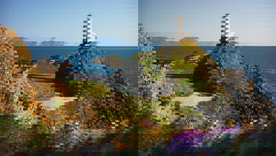 The Twr Mawr Lighthouse placed upon a green hill on a sandy beach, with mountains in the background