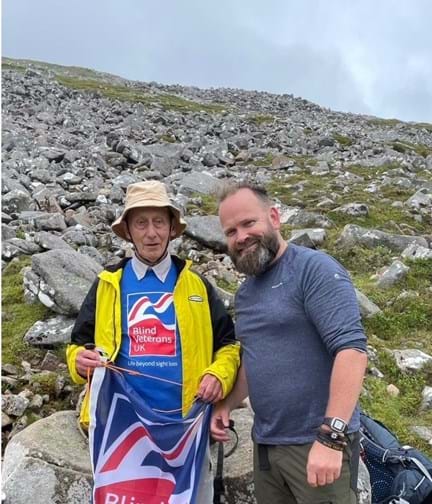 Volunteer Chris and blind veteran Pete pictured at the highest point reached on Ben Nevis, waving a Blind Veterans UK flag