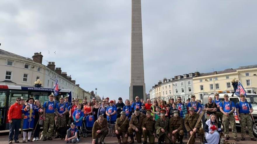 Group of walkers, some in Blind Veterans UK T-shirts, some in military themed costumes, gathered in front of Llandudno War Memorial.
