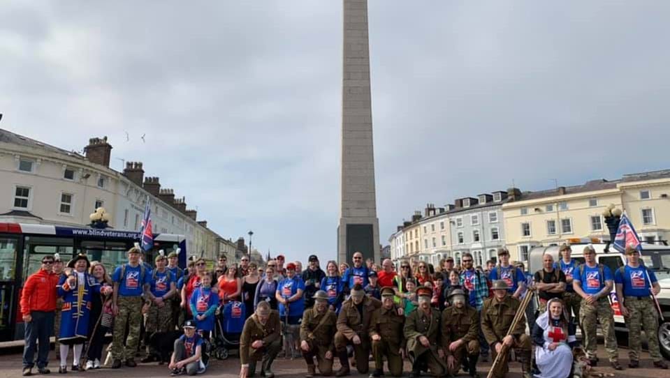Group of walkers, some in Blind Veterans UK T-shirts, some in military themed costumes, gathered in front of Llandudno War Memorial.