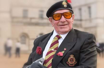 A smiling blind veteran wearing large orange tint glasses, a beret, with military medals and poppy pinned to his jacket.