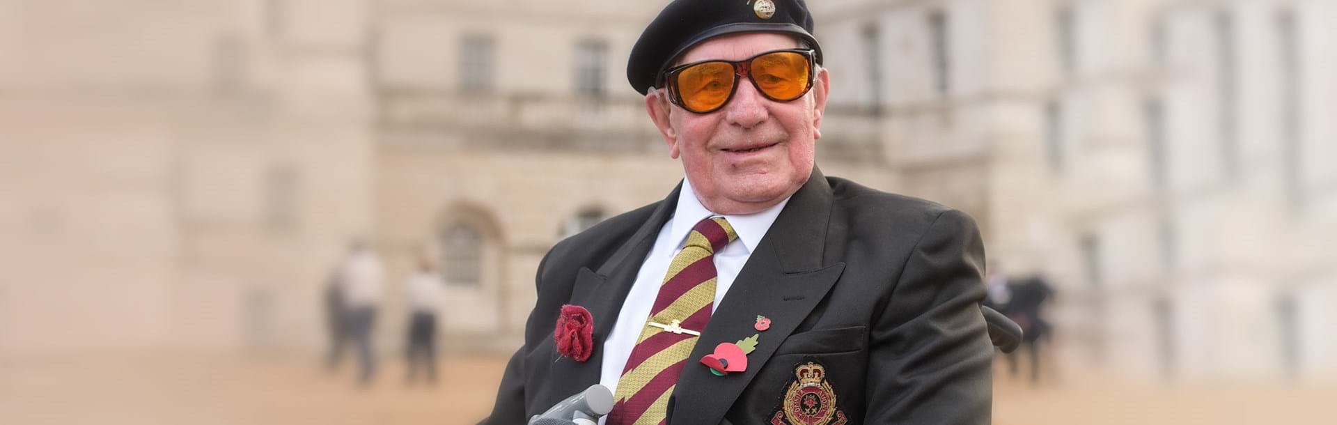 A smiling blind veteran wearing large orange tint glasses, a beret, with military medals and poppy pinned to his jacket.