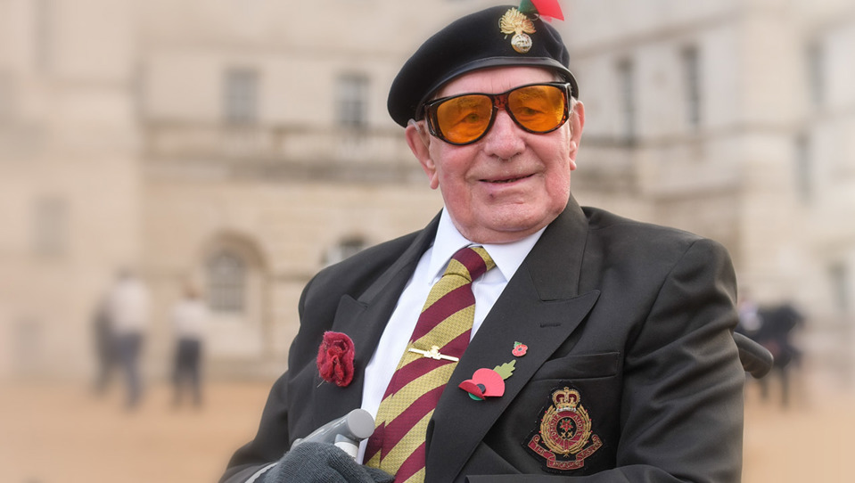 A smiling blind veteran wearing large orange tint glasses, a beret, with military medals and poppy pinned to his jacket.