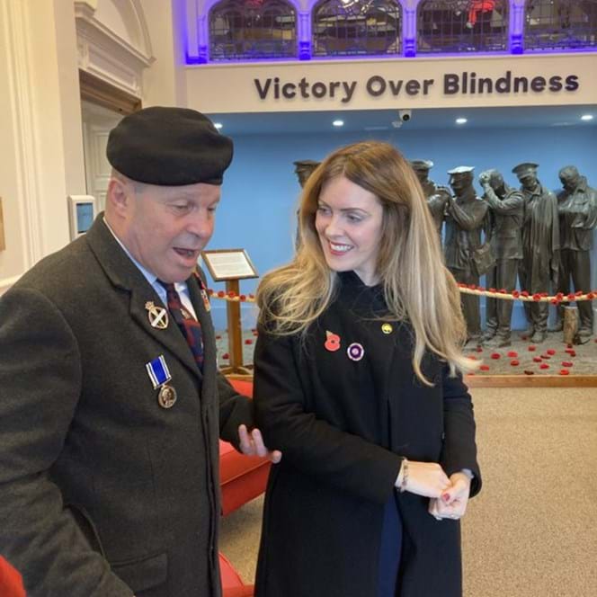 Billy wearing his medals is stood holding Kelly's arm in front of the Victory of Blindness sculpture
