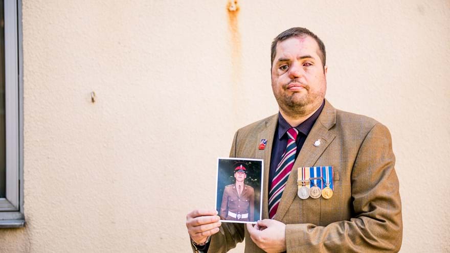 Blind veteran Simon is wearing a blazer, tie, and medals. He looks into camera and holds a photo of himself with both hands.