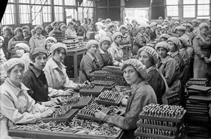 A large group of women munition workers in a factory, sitting at long tables with bullets and casings heaped on them