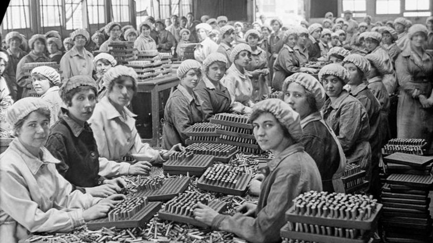 A large group of women munition workers in a factory, sitting at long tables with bullets and casings heaped on them