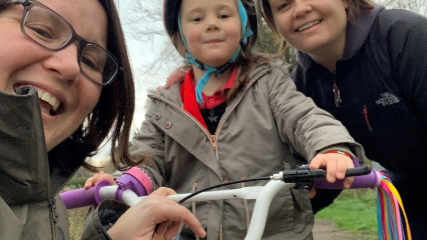 Photo of Kelly Ganfield, right, smiling with wife Sarah, left, and young daughter Bethany on trike, centre