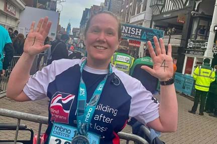 A photo of Alison wearing her medal and a Blind Veterans UK vest after finishing the Chester 10K, raising her hands to show her palms, which show the numbers 1 and 4