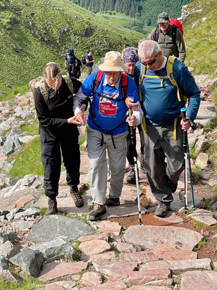 Blind veteran Peter being guided and supported up a rocky part of the mountain by his family and fellow climbers