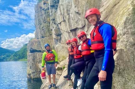 Blind veteran Jules, joined by other blind veterans and guides, wearing life jackets and we suits, smiling as he leans against a rocky wall on the edge of a lake,