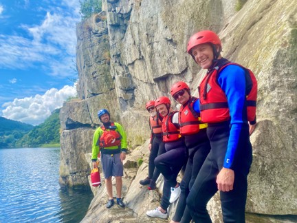 Blind veteran Jules, joined by other blind veterans and guides, wearing life jackets and we suits, smiling as he leans against a rocky wall on the edge of a lake,