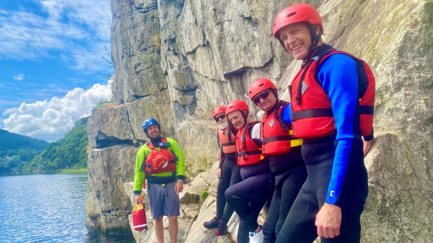 Blind veteran Jules, joined by other blind veterans and guides, wearing life jackets and we suits, smiling as he leans against a rocky wall on the edge of a lake,