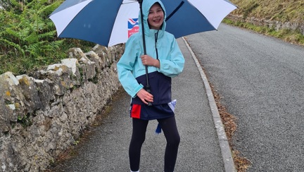 A photo of Emmie, wearing a a hooded rain coat and holding a Blind Veterans UK umbrella, walking along a curved path 