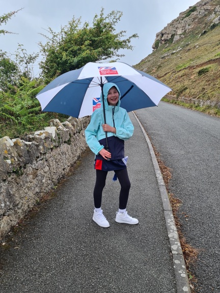 A photo of Emmie, wearing a a hooded rain coat and holding a Blind Veterans UK umbrella, walking along a curved path
