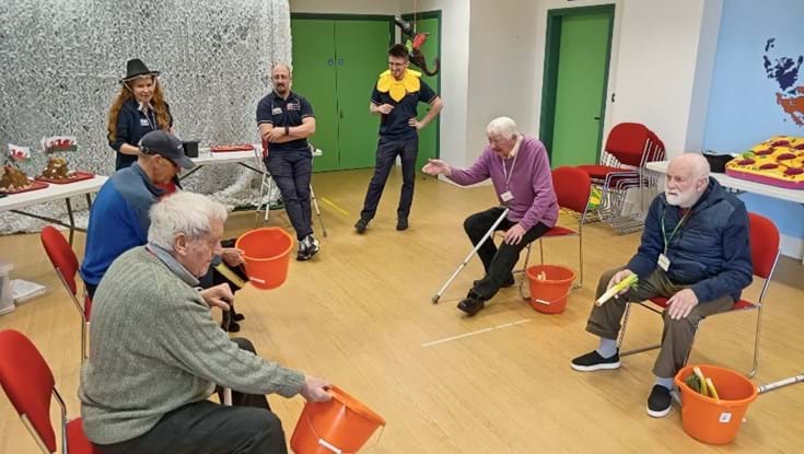 A group of people sit on chairs in a circle holding buckets and attempt to throw leeks into the buckets
