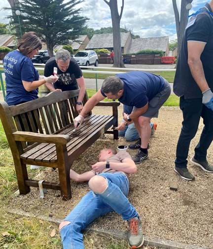 Four Thea colleagues working together to varnish a wooden garden bench at our Blind Veterans UK Llandudno Centre of Wellbeing.  One of the group is lay on the floor so he can reach underneath.