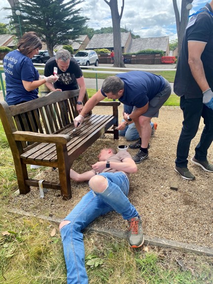 Four Thea colleagues working together to varnish a wooden garden bench at our Blind Veterans UK Llandudno Centre of Wellbeing. One of the group is lay on the floor so he can reach underneath.