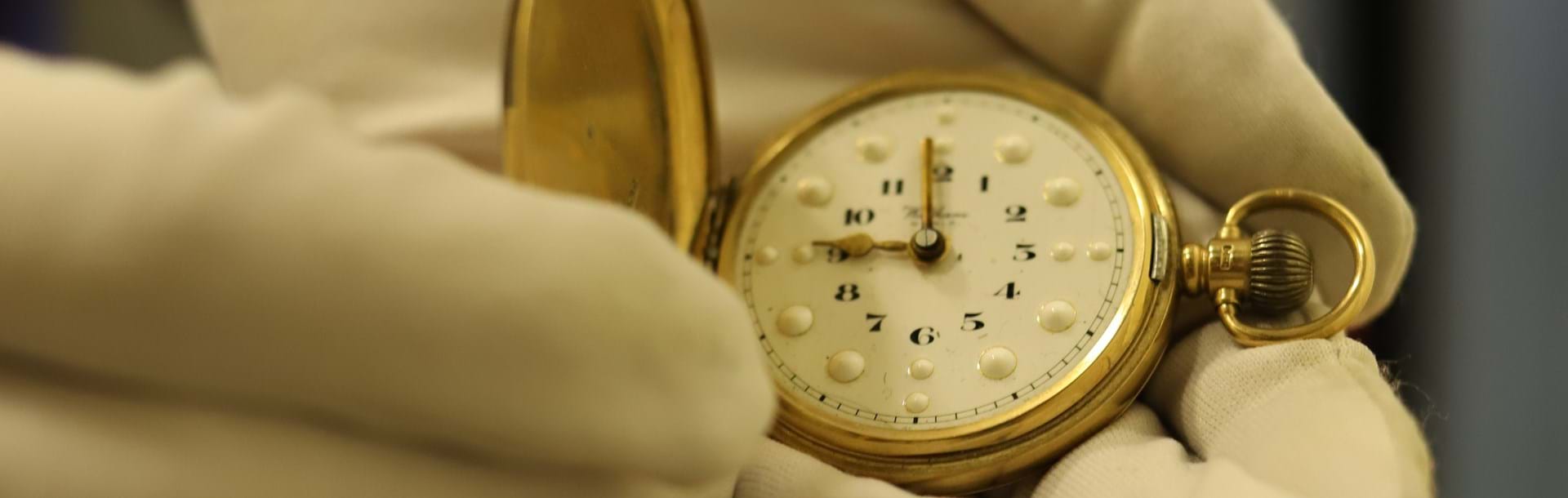 An open gold braille pocket watch, being held by a pair of hands wearing white cotton gloves