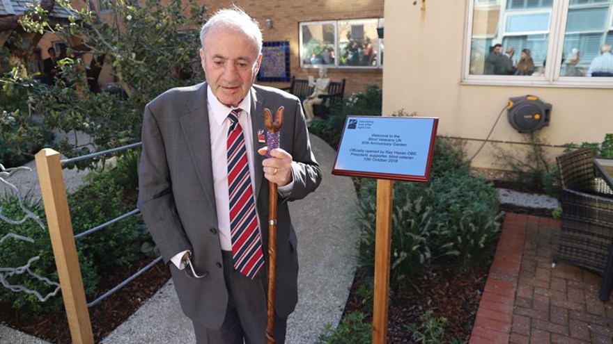 Blind veteran Ray Hazan OBE, holding a walking stick, standing in the garden of our centre of wellbeing, next to a plaque commemorating the opening of the garden.