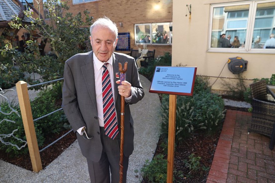 Blind veteran Ray Hazan OBE, holding a walking stick, standing in the garden of our centre of wellbeing, next to a plaque commemorating the opening of the garden.
